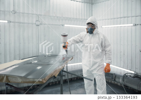 A worker dressed in a protective suit is spray-painting a car panel inside a modern paint booth 133364193