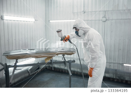 Automotive technician spray-painting car panel in protective suit inside paint booth 133364195