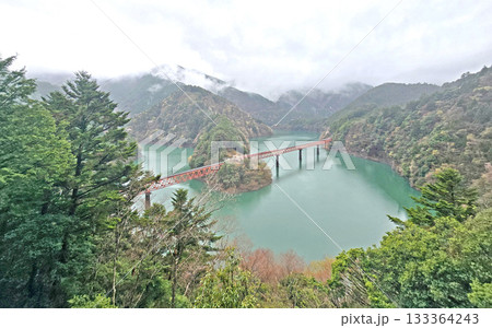 Scenic view of a red bridge over a lake surrounded by lush green mountains Scenic view of a red bridge over a lake surrounded by lush green mountains 133364243
