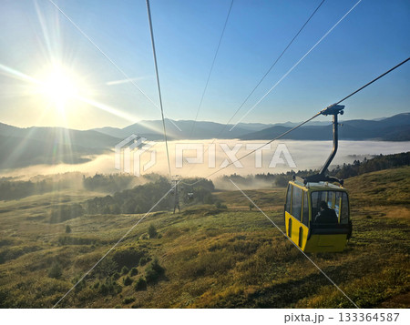 A yellow cable car ascends a mountain, offering stunning views of the valley and the morning sun A yellow cable car ascends a mountain, offering stunning views of the valley and the morning sun 133364587