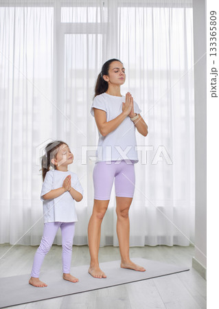 Mother and daughter white and purple sportswear standing in prayer pose on a yoga mat 133365809