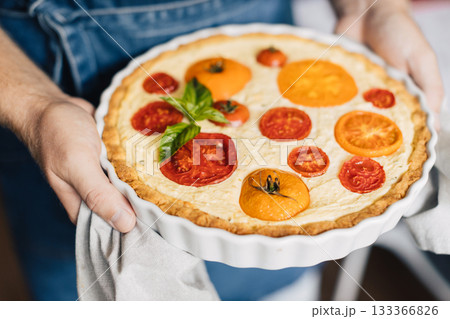 Man hands holding cheese and tomato quiche in the ceramic baking dish. Man hands holding cheese and tomato quiche in the ceramic baking dish. 133366826