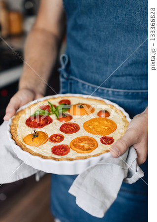 Man hands holding cheese and tomato quiche in the ceramic baking dish. 133366828