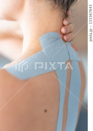 Close up view of young woman with kinesiology tape applied to her neck and shoulder holding her neck with her hand. Physical therapy and muscle recovery through modern rehabilitative care 133367643