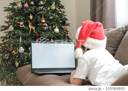 Boy in Santa hat using laptop with blank screen on sofa next to decorated Christmas tree. Concept: holidays, video calls, online shopping, remote learning, screen mockup 133369893