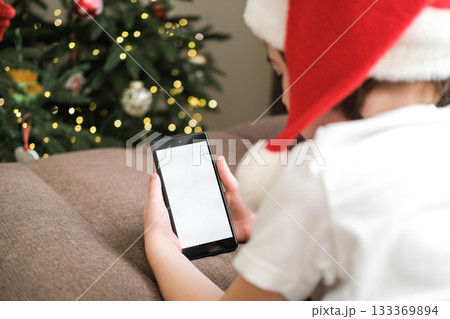 Five-year-old boy in Santa hat using a smartphone with blank white screen on sofa next to decorated Christmas tree. Christmas holidays, video calls, mobile apps, screen mockup Five-year-old boy in Santa hat using a smartphone with blank white screen on sofa next to decorated Christmas tree. Christmas holidays, video calls, mobile apps, screen mockup 133369894