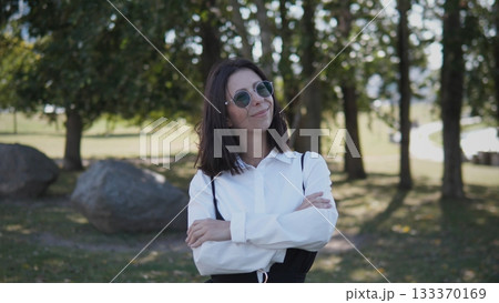 A businesswoman stands confidently in a sunny park, dressed in a white blouse and sunglasses. She smiles while crossing her arms, surrounded by trees and large stones. A businesswoman stands confidently in a sunny park, dressed in a white blouse and sunglasses. She smiles while crossing her arms, surrounded by trees and large stones. 133370169