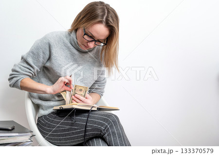 blond woman with glasses counting money and writing in a notebook, isolated on a gray 133370579
