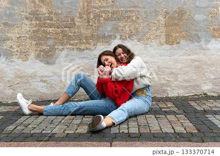 two young women hugged sitting on a paved sidewalk by an old wall 133370714