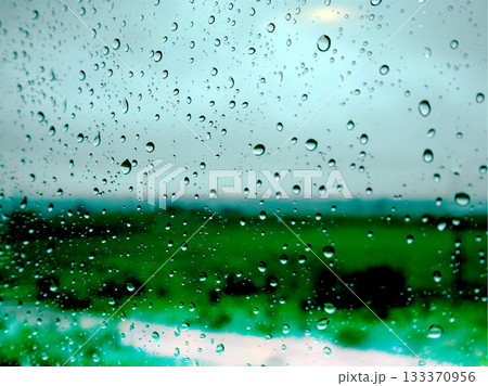 Raindrops on glass with a blurred green field and gloomy grey sky in the background.Water droplets on a window in rainy season. Raindrops on glass with a blurred green field and gloomy grey sky in the background.Water droplets on a window in rainy season. 133370956