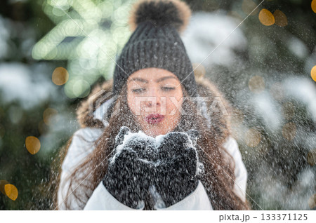 young woman blows snow from her hands while standing against the background of a Christmas tree 133371125