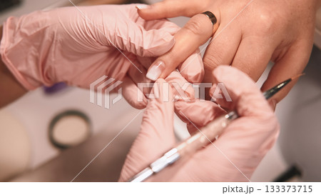 Nail care. Nude polish. Beautician in latex gloves working on female client hand using professional tool acrylic gel manicure in pastel colors. Nail care. Nude polish. Beautician in latex gloves working on female client hand using professional tool acrylic gel manicure in pastel colors. 133373715