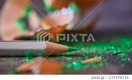 A green pencil rests next to wood shavings on a surface. A green pencil rests next to wood shavings on a surface. 133376216