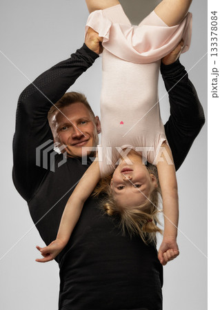 little blonde girl is hanging upside down while her father holds her playfully in a studio setting. Both are smiling and enjoying their time together sharing a joyful moment of laughter. 133378084