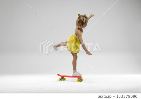 small blonde girl practices her balance on a skateboard in a studio. She wears a bright yellow dress and shows confidence while having fun. The simple background highlights her joyful activity. 133378090