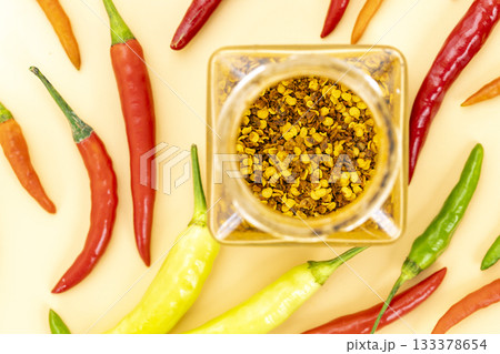 Top view of crushed red chili flakes in a glass jar with fresh colorful red, green, yellow, and orange chili peppers scattered Top view of crushed red chili flakes in a glass jar with fresh colorful red, green, yellow, and orange chili peppers scattered 133378654