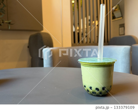 Creamy matcha bubble tea in a sealed plastic cup sits on a cafe table, with soft indoor lighting and a neutral modern interior in the background 133379109