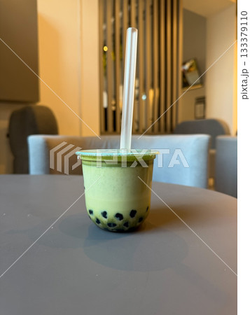 Creamy matcha bubble tea in a sealed plastic cup sits on a cafe table, with soft indoor lighting and a neutral modern interior in the background 133379110