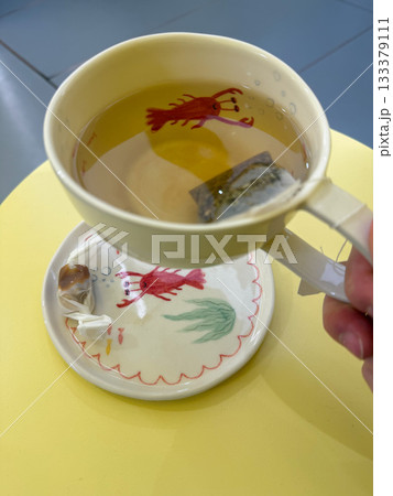 A ceramic cup of hot tea with a lobster illustration inside sits on a matching saucer with playful marine motifs, set on a pastel yellow table 133379111