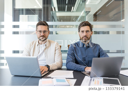 Two professional businessmen sitting at a modern office desk, actively working on their laptops and papers while looking directly at the camera during a collaborative corporate meeting Two professional businessmen sitting at a modern office desk, actively working on their laptops and papers while looking directly at the camera during a collaborative corporate meeting 133379141