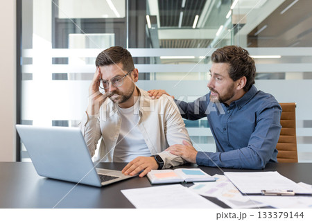 Two male coworkers in a modern office, one offering comfort and support to the other who is stressed, holding his head over a laptop amid paperwork and tension 133379144