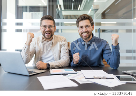 Two cheerful businessmen sitting at a desk with laptops and documents, raising their fists in celebration, happy about achieving a goal or closing a successful deal 133379145