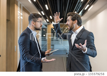Two male colleagues arguing intensely in a modern open-plan office, shouting and gesturing in a heated professional dispute while one holds a tablet, showing stress and tension 133379258