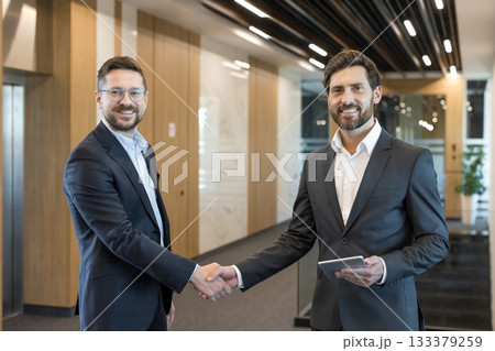 Two smiling businessmen in suits are shaking hands, confirming a successful partnership and agreement in a modern office hallway, one holding a digital tablet 133379259