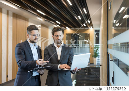 Two professional businessmen discussing project details in a modern corporate office corridor, one holding a laptop while the other points to a clipboard, representing teamwork and efficiency 133379260