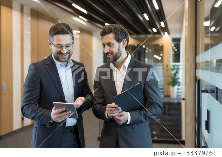 Two professional businessmen smiling and looking at a digital tablet, sharing ideas and discussing work while walking through a contemporary office hallway Two professional businessmen smiling and looking at a digital tablet, sharing ideas and discussing work while walking through a contemporary office hallway 133379261