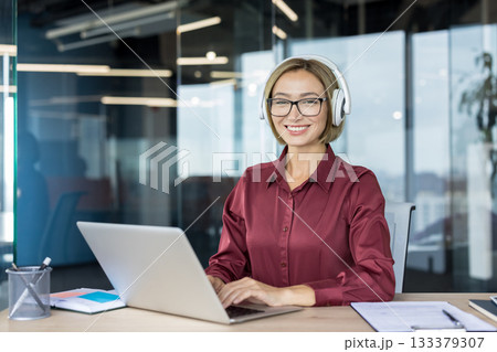Smiling businesswoman wearing glasses and white headphones, sitting at an office desk and happily working on a laptop, embodying professionalism, concentration, and modern workplace communication 133379307
