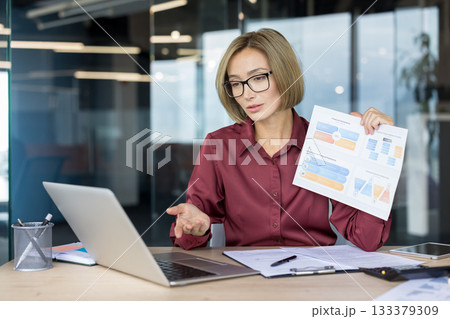 Businesswoman discussing business strategy and financial data, holding a document with graphs during a remote video conference meeting using her laptop in a modern office Businesswoman discussing business strategy and financial data, holding a document with graphs during a remote video conference meeting using her laptop in a modern office 133379309