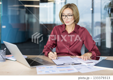 Professional businesswoman in glasses calculating expenses and writing notes on documents, managing finance and accounting tasks at a modern office desk with laptop and charts 133379313