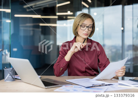 Businesswoman reviewing financial reports and data on paper, concentrating on her work at a desk with a laptop and various documents in a contemporary office environment Businesswoman reviewing financial reports and data on paper, concentrating on her work at a desk with a laptop and various documents in a contemporary office environment 133379316
