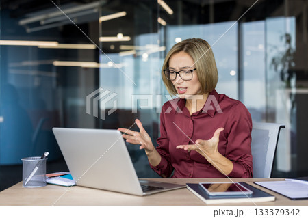 Businesswoman feeling frustrated and stressed, gesturing with her hands while having a challenging discussion or problem-solving during a video conference on her laptop in a modern office 133379342