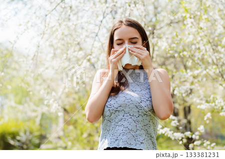 Sneezing young girl with handkerchief among blooming trees in spring park. Concept of allergy Sneezing young girl with handkerchief among blooming trees in spring park. Concept of allergy 133381231