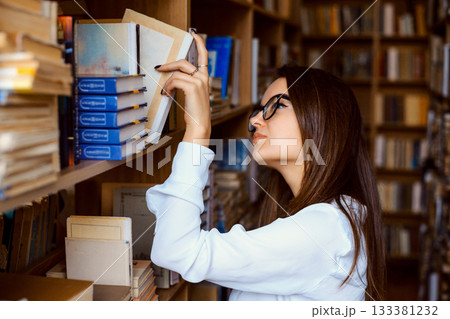 Female college student taking book from shelf in library. Hard-working student looking for a book to find necessary information and prepare report of historical event 133381232