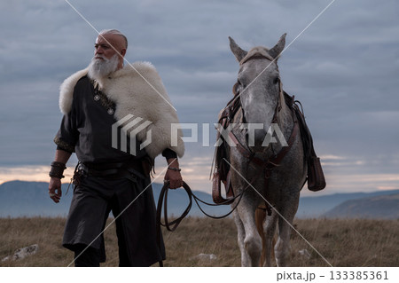 A majestic Viking chieftain on a dappled grey horse, clad in a fur cloak, surveys a desolate, mountainous landscape under a dramatic cloudy sky, exuding power and presence. 133385361
