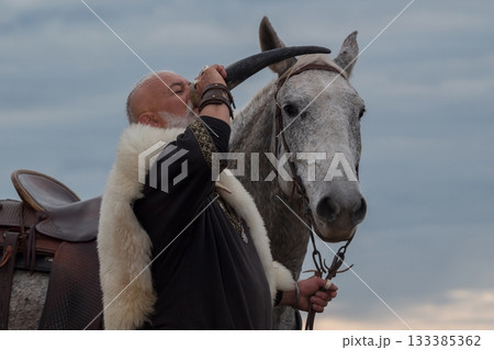 A rugged Viking man with a white beard, dressed in traditional attire, drinking from a horn against a stunning sunset background, showcasing strength, heritage, and heroic spirit 133385362