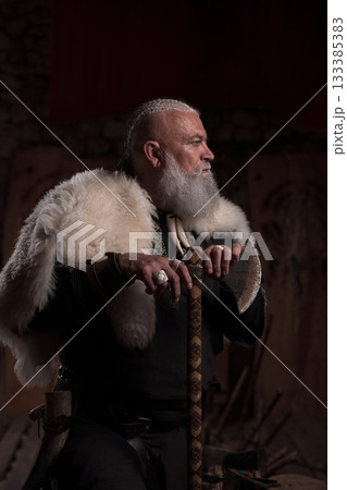 A rugged Viking man with a white beard, wearing traditional armor and a fur cloak, stands inside a stone hall, holding a sword with a contemplative expression, evoking strength and heritage. 133385383