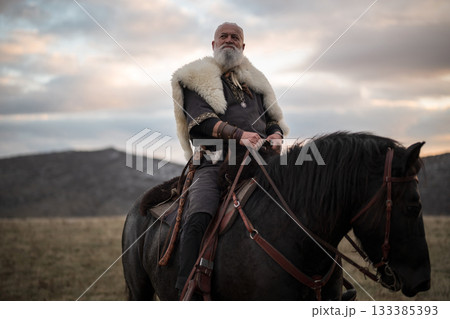 A Viking warrior on a large black horse surveys a dramatic landscape during sunset, wearing a fur cloak. The scene evokes ancient exploration, adventure, and the spirit of the wild. 133385393