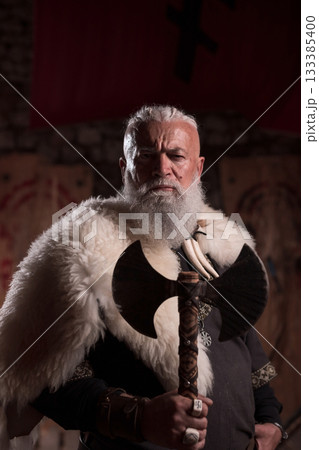Rugged Viking warrior as a king or hero with a white beard, dressed in traditional armor, holding a battle axe, inside a medieval hall, intense and fierce, embodying strength and heritage 133385400