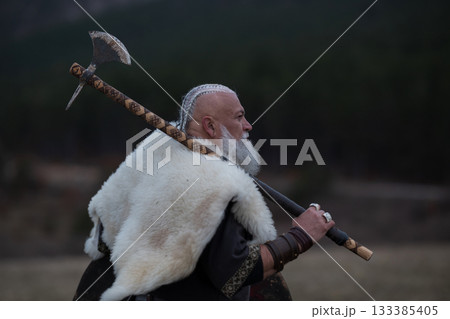 Viking warrior with axe standing on mountain cliff, wearing fur cloak and leather armor, overlooking dramatic landscape. Historical Nordic fighter 133385405