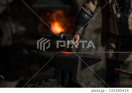 A Viking blacksmith with a white beard and braided hair works intently at a forge, hammering glowing metal on an anvil, with flames flickering in the background of a stone workshop. 133385414