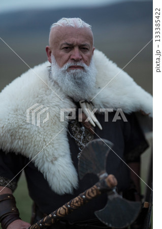 Viking warrior with axe standing on mountain cliff, wearing fur cloak and leather armor, overlooking dramatic landscape. Historical Nordic fighter 133385422