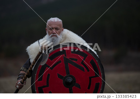 Viking warrior with axe standing on mountain cliff, wearing fur cloak and leather armor, overlooking dramatic landscape. Historical Nordic fighter 133385423