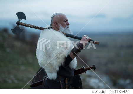 Viking warrior with axe standing on mountain cliff, wearing fur cloak and leather armor, overlooking dramatic landscape. Historical Nordic fighter 133385424