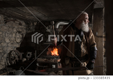 A Viking blacksmith with a white beard and braided hair works intently at a forge, hammering glowing metal on an anvil, with flames flickering in the background of a stone workshop. 133385435