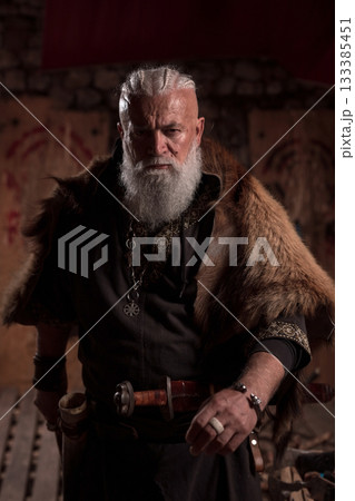 Elderly Viking man with a white beard in traditional attire, wielding a sword, exuding confidence and strength in a historic, dimly-lit setting 133385451