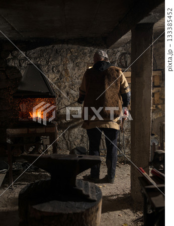 A Viking blacksmith with a white beard and braided hair works intently at a forge, hammering glowing metal on an anvil, with flames flickering in the background of a stone workshop. 133385452